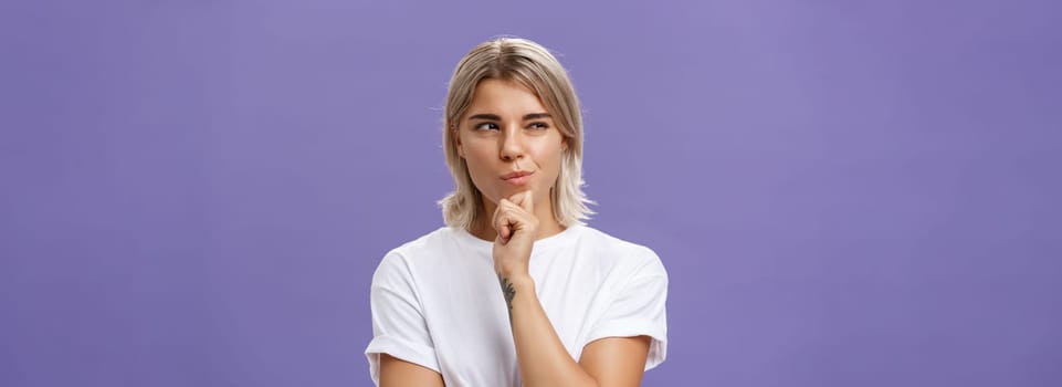 Waist-up shot of smart creative young ambitious woman with blond haircut and tanned skin squinting and smirking gazing left while holding hand on chin thinking making up plan or decision. Copy space