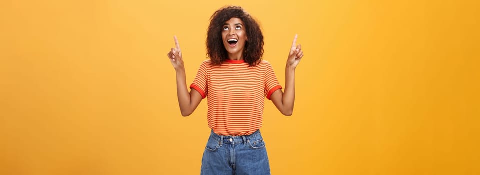 Fascinated impressed and amused good-looking charming African-American woman with afro hairstyle in trendy t-shirt and denim shorts pointing and looking up with interested look over orange wall. Lifestyle.
