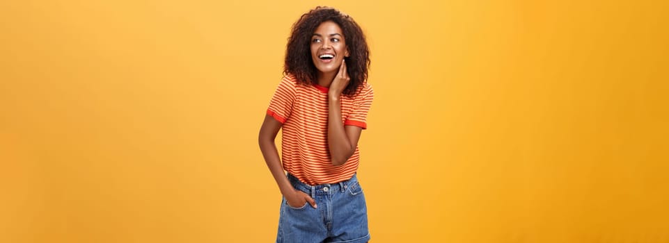 Stylish feminine and fashionable african american female model with afro hairstyle touching neck gently looking right with amused carefree expression holding hand in pocket over orange background. Lifestyle.