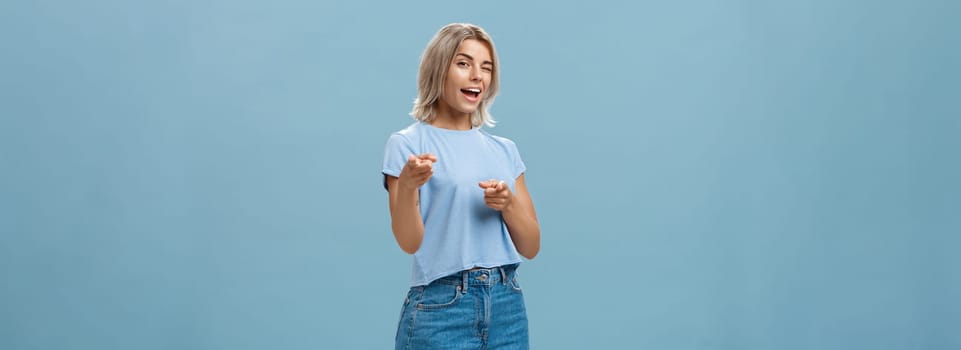 Hey I got offer for you. Portrait of attractive self-assured and playful stylish blond female with tanned skin in denim shorts and summer t-shirt winking and pointing at camera over blue background.