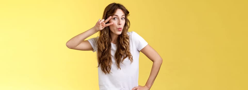 Cute glamour young woman curly haircut tilt head silly show peace victory sign fold lips lovely muah kiss gesture send flirty glances camera hold hip mimicking sensually gazing you yellow background. Lifestyle.