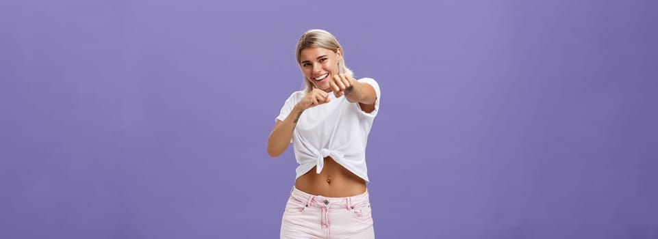 Ready fight back. Energized happy and awesome blonde female with tanned skin and fit body pulling fist in attack gesture towards camera smiling joyful acting like fighting or boxing over purple wall.