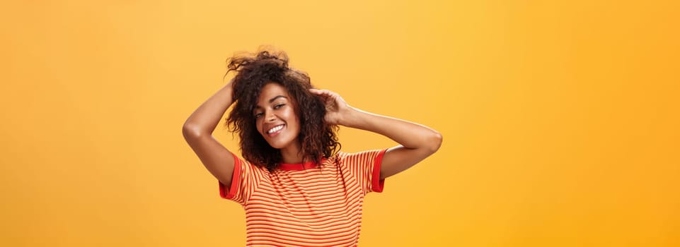 Waist-up shot of charming flirty feminine dark-skinned female in playful mood dancing playing with curly hair and smiling with delight and joy posing over orange background happy and carefree. Lifestyle.
