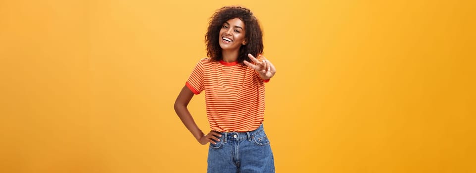 Friendly carefree and happy good-looking tall african american female model with curly hairstyle holding hand on hip tilting head and smiling joyfully showing peace gesture over orange wall. Copy space