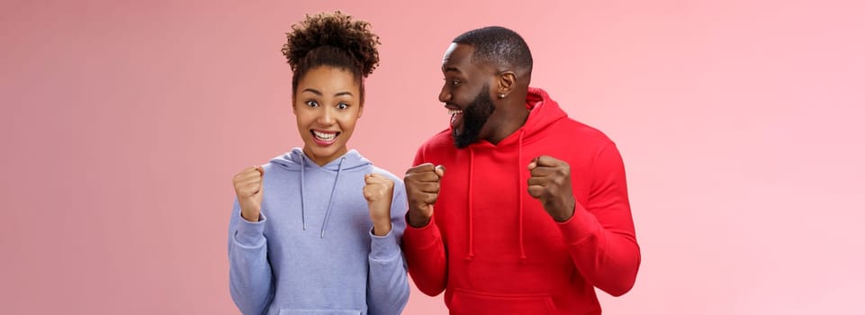 Happy enthusiastic thrilled young african american couple clenching fists joyfully surprised winning awesome honeymoon trip tickets standing pink background triumphing smiling broadly celebrating.