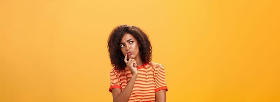 Woman considering to take part in event. Smart focused and creative good-looking african girl with curly hair holding finger on lip gazing concerned left, thinking making decision over orange wall.