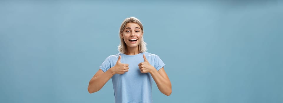 You did great proud of you. Portrait of satisfied and fascinated attractive happy girlfriend in casual t-shirt showing thumbs up and smiling broadly being supportive and cheerful over blue wall.