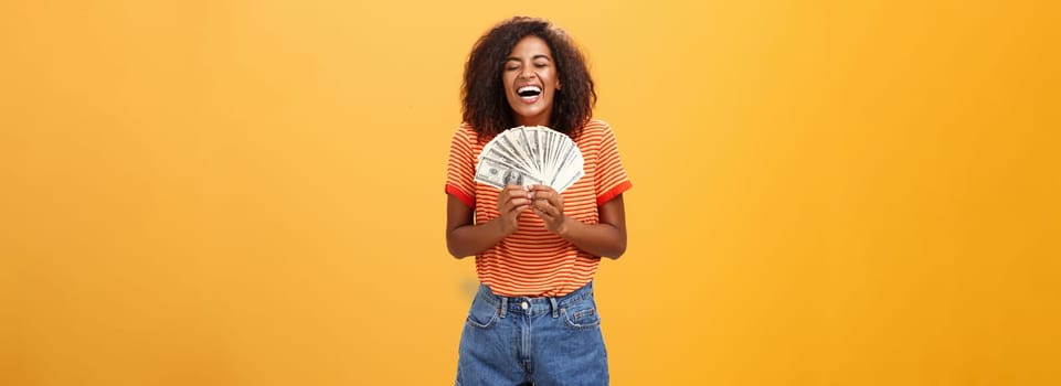 Finally girl rich. Portrait of happy charismatic stylish young female model receiving first payment holding lots of money and laughing from joy winning lottery ready waste cash over orange background.