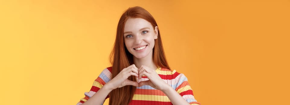 Lovely friendly-looking charismatic smiling redhead girl express affection love and friendship grinning show heart sign chest showing sympathy, confess romantic feelings, standing orange background.