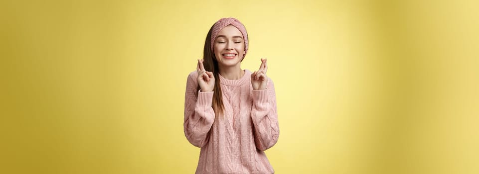 Girl wishing wellness wanting dream come true, cross fingers dreamy, close eyes waiting miracle, anticipating good news, posing excited and joyful against yellow background in knitted warm sweater.