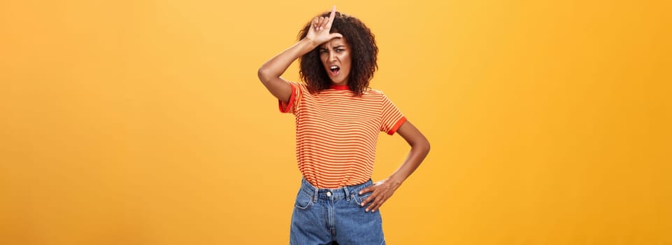 Arrogant and confident woman not talk with losers. Mocking self-assured stylish african american female with curly hair in trendy clothes frowning scolding nerd, showing letter l on forehead. Copy space