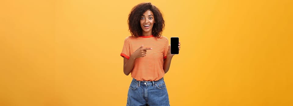 Cute african american stylish woman showing friend new app in smartphone pointing at cellphone screen with index finger smiling thrilled and happy delighted with cool device purchase over orange wall. Lifestyle.