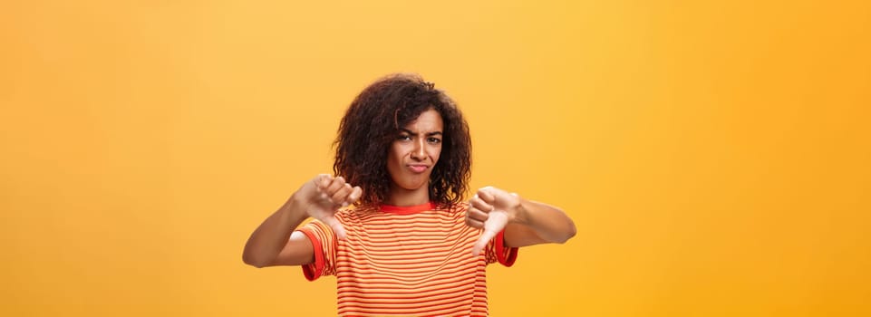 You loser and idea sucks. Dissatisfied and displeased young creative female artist with curly hairstyle frowning, sulking showing thumbd down in dislike gesture giving negative answer over orange wall.