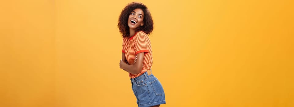 Hey wanna hang out. Sensual and flirty carefree stylish dark-skinned woman with curly hair standing in profile turning left and posing with happy cute expression smiling and seducing over orange wall.