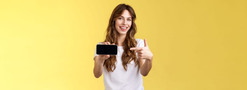 Confident sassy good-looking stylish woman introduce awesome game app show smartphone pointing index finger blank mobile phone screen smiling delighted recommend try application yellow background. Lifestyle.