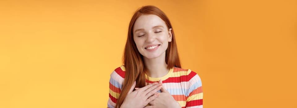 Happy romantic delighted sighing young lovely redhead girlfriend smiling broadly touching heart chest palms close eyes recalling date passionate kiss feel gratitude warmth, orange background.