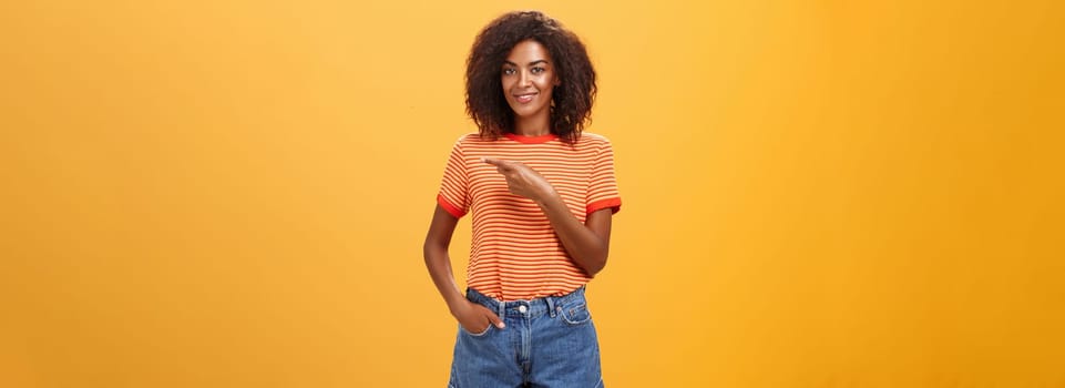 Showing perfect place for advertising. Charming carefree and confident young stylish african-american woman with afro hairstyle holding hand in pocket casually pointing right over orange wall.