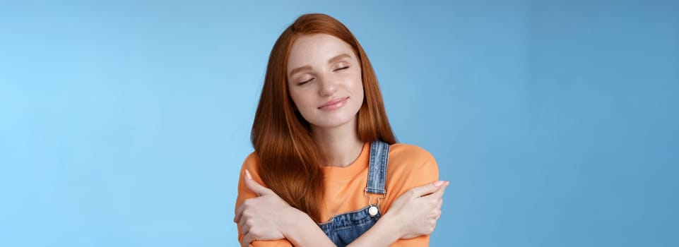 Passionate romantic tender ginger girl feel safe comfort close eyes smiling gently lovely daydreaming hugging herself recalling boyfriend cuddles sensual embraces, standing blue background happy.