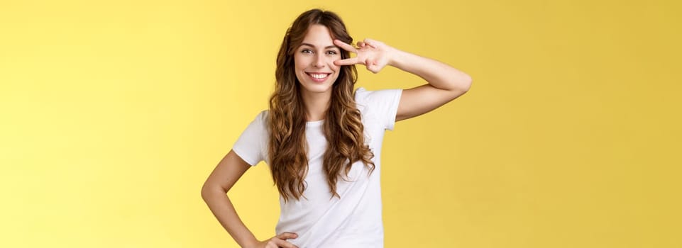 Cheerful positive cute girl curly-haired show peace sign delighted enjoy youth having perfect summer holidays determined rest trip vacation smiling broadly stand confident yellow background. Lifestyle.