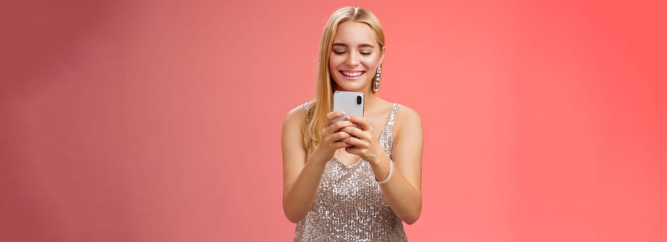 Delighted tender glamour blond woman in silver stylish glittering dress brilliand earrings holding smartphone taking photo friend capture moment celebration nightclub, red background smiling.