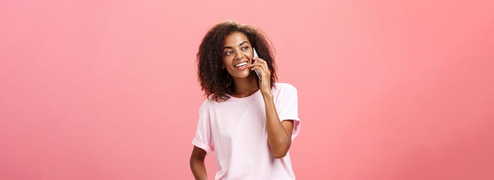 Girl calling friend to meet up. Portrait of charming friendly and outgoing african american young woman with afro hairstyle holding smartphone near ear while talking looking left curiously. Copy space