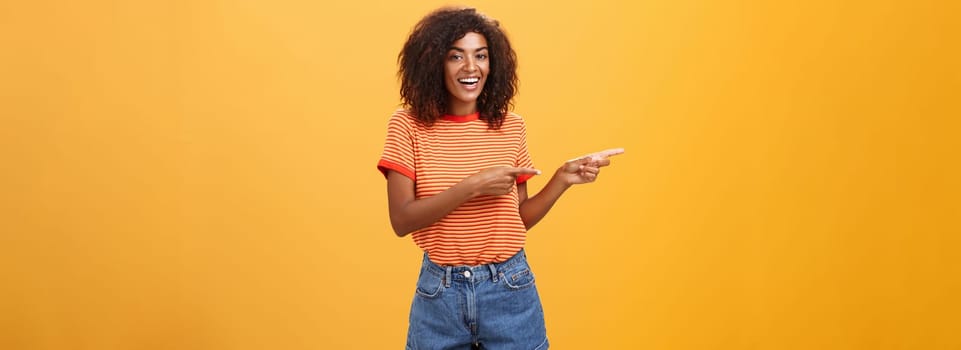 Ask him. Portrait of friendly and joyful good-looking stylish female shop assistant with curly hair and dark skin pointing left with both hands, smiling assured and entertained over orange background.