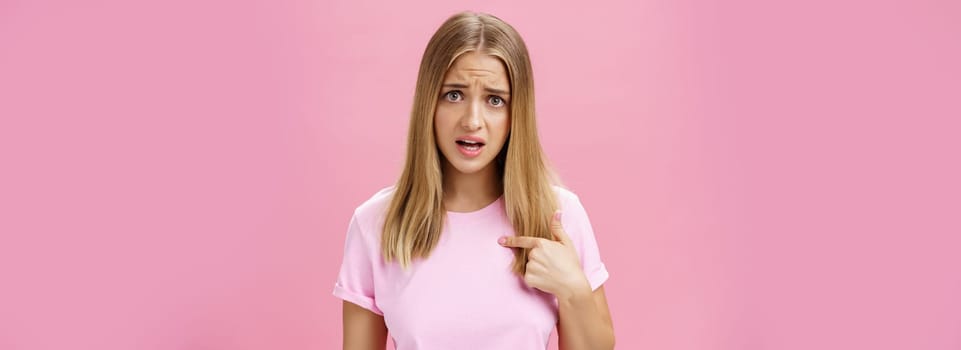 Insulted girl pointing at herself with displeased pissed and questioned expression asking question being shocked she picked or accused in something terrible and disrespectful posing against pink wall. Lifestyle.