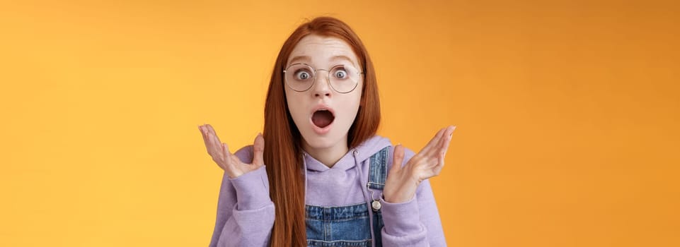 Close-up shocked sensitive concerned young panicking redhead woman worry drop jaw gasping raise hands spread freak out stare surprised emotional reacting incredible news, orange background.