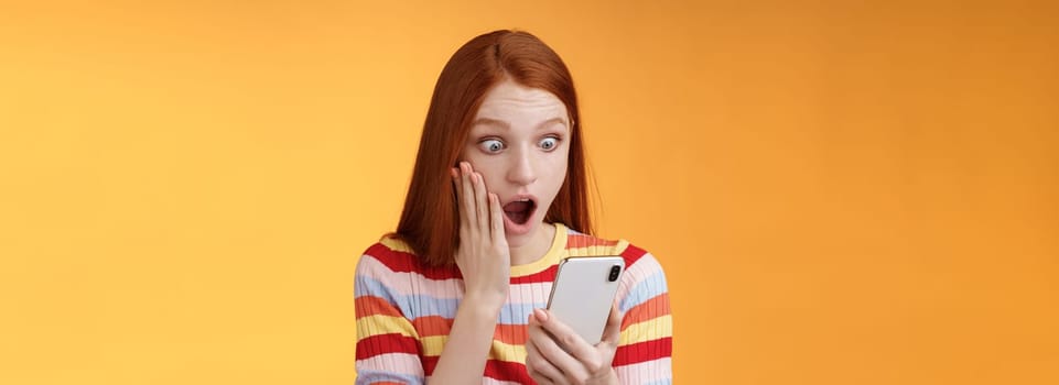 Omg what heck. Portrait shocked concerned young redhead sensitive impressed redhead woman stare smartphone display touch cheek drop jaw stunned surprised standing orange background hold phone.