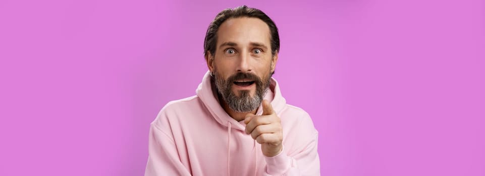 Fascinated amused european bearded mature guy widen eyes shocked open mouth recognize person pointing camera index finger surprised see familiar famous star, standing purple background.