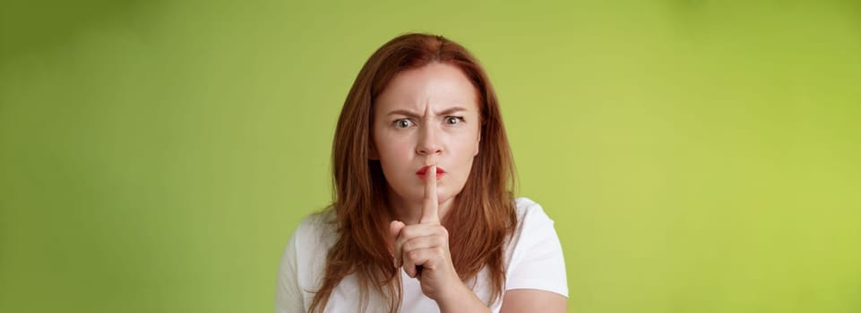 Not speak during exam. Strict serious-looking displeased middle-aged redhead woman frowning disappointed hushing say shush index finger pressed lips keep quiet gesture green background.