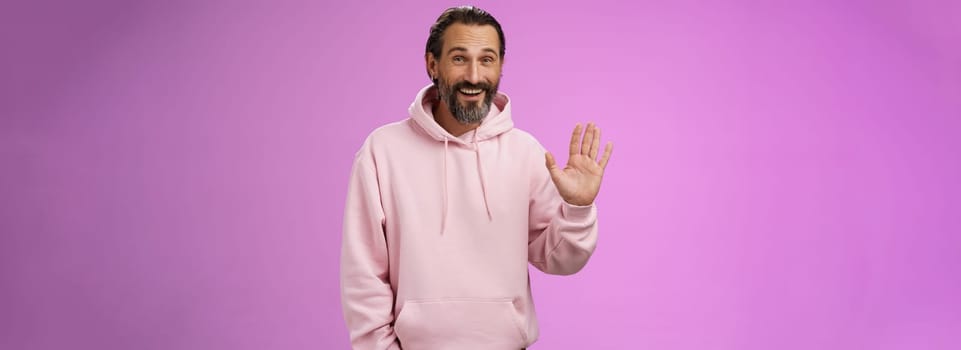 Cheeky charismatic funny happy smiling mature man bearded grey hair in pink hoodie waving palm hello nice meet you greetng gesture welcoming guests say hi standing purple background friendly relaxed. Lifestyle.