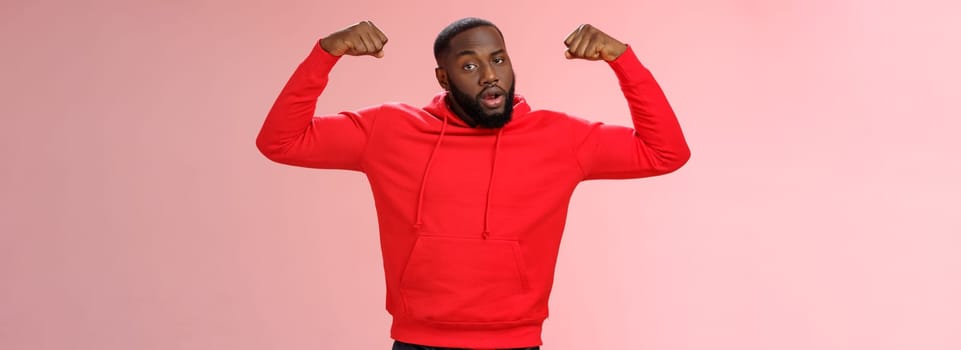Strong cheeky serious-looking african american sportsman in red hoodie bragging showing-off raise hands excercising show big muscles biceps, standing confident pink background. Copy space