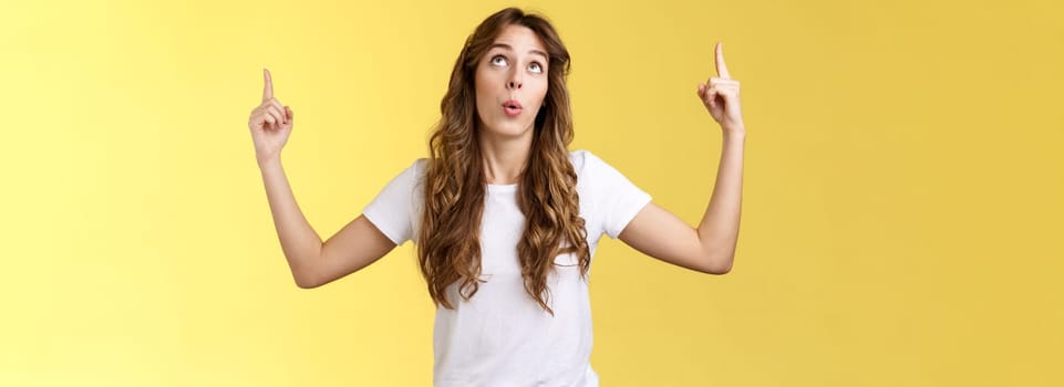 Impressed curious wondered attractive european woman long curly haircut look pointing index fingers up top copy space promo react astonished fascinated surprising event yellow background.