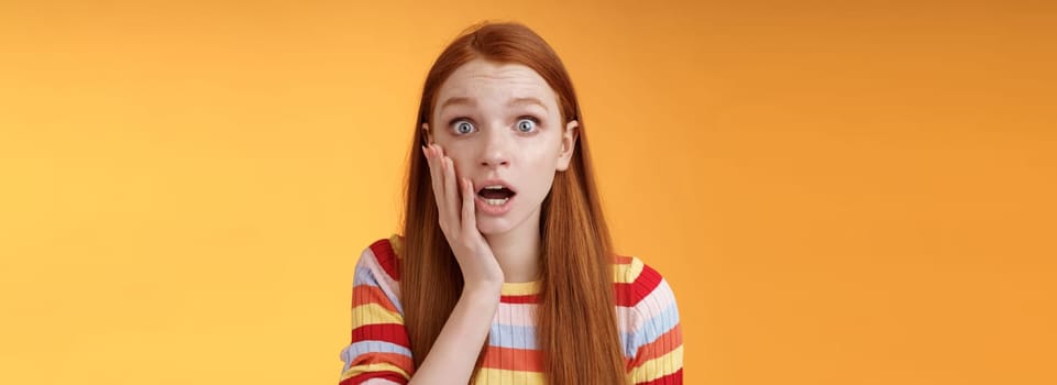 Concerned worried ginger girl blue eyes drop jaw gasping touch cheek confused looking nervously anxious, show empathy hearing terrible upsetting story standing orange background. Copy space