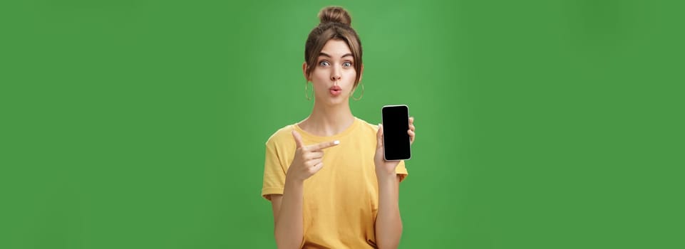 Portrait of intrigued woman cannot wait use new smartphone holding phone and pointing at cellphone screen folding lips with excitement and interest presenting new device model in store over green wall. Lifestyle.