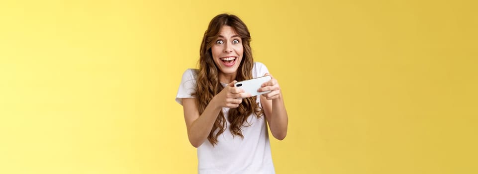 Happy enthusiastic excited cute playful girl winning awesome smartphone game thrilled playing stare camera surprised triumphing smiling happily hold mobile phone horizontal yellow background.