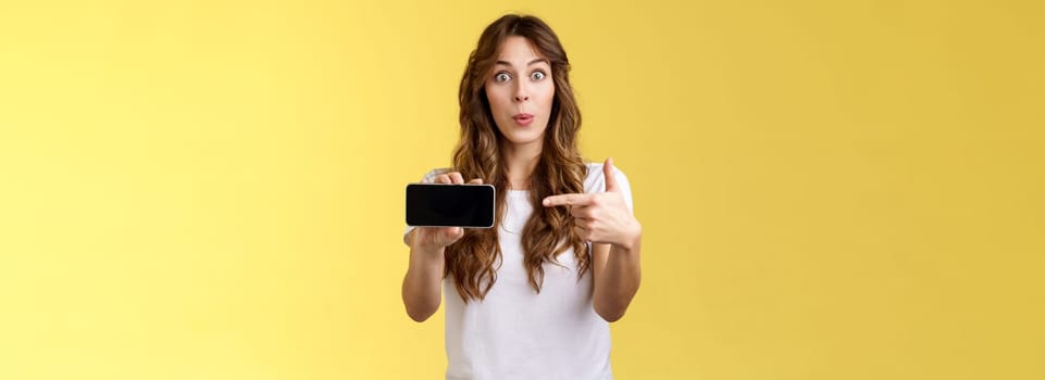 Excited surprised pleased girl curly long hairstyle folding lips whistling amused stare camera impressed showing smartphone pointing index finger mobile cellphone screen stand yellow background.