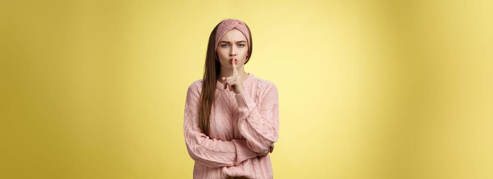 Keep mouth shut. Serious-looking bossy attractive young 20s woman in sweater, headband shushing making shhh gesture holding index finger on lips, gossiping, spread rumors over yellow background.