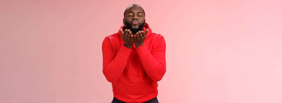 Charming cute african american bearded guy in red hoodie close eyes folding lips hold palms near mouth sending passionate air kiss blowing mwah camera, standing lovely pink background.