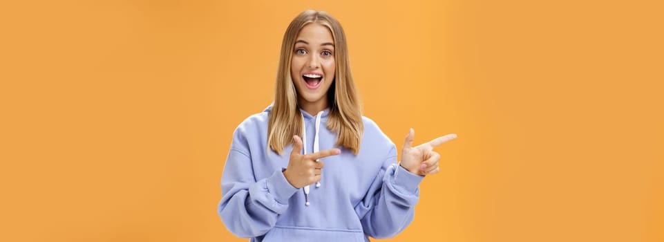 Amused girl pointing at curious copy space showing left with index fingers smiling excited and surprised standing entertained with upbeat grin in cozy blue hoodie over orange background. Lifestyle.