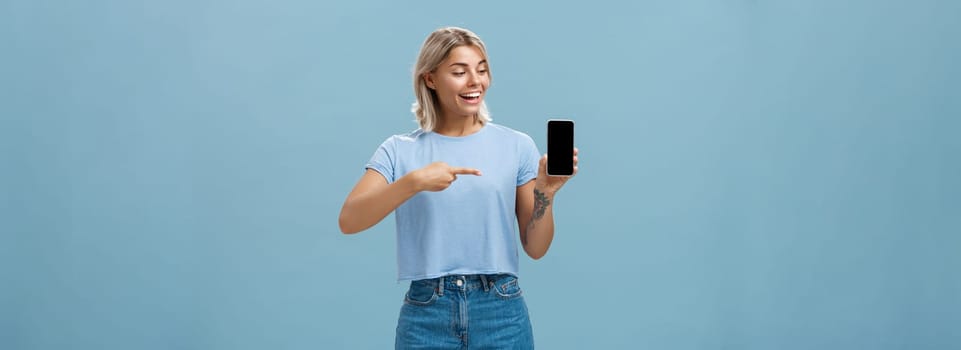 Portrait of delighted good-looking european female with blond haircut showing smartphone screen with amazement and delight pointing at device while promoting brand new gadget over blue background. Technology concept