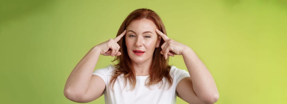 Smart choice. Intrigued thoughtful good-looking assertive redhead creative middle-aged woman touch temples squinting pondering info thinking use mental power read minds stand green background.