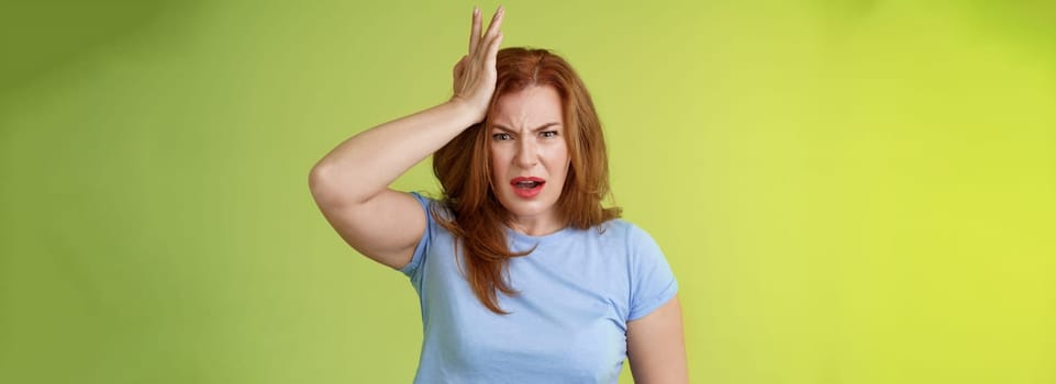 Ouch you kick my head. Frustrated concerned upset middle-aged redhead woman look shocked distressed touch temple complain kid shoot ball her face stand disappointed green background. Copy space
