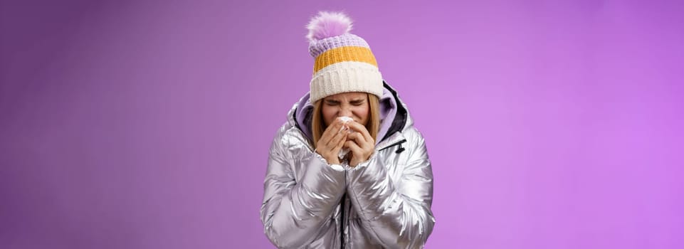 Girl runny nose sneeze tissue press napkin face got ill feeling unwell sick heading hospital standing purple background bending wearing silver stylish jacket winter hat, crying sobbing.