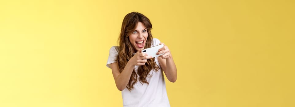Excited intense playful gamer girl geek playing awesome smartphone game hold mobile phone horizontal grimacing eager win arcade stare screen tap display stand yellow background tilting. Lifestyle.