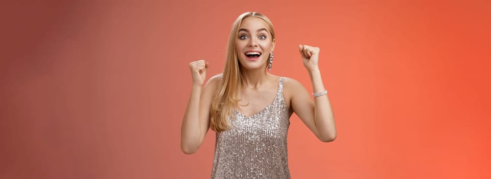 Lifestyle. Surprised happy celebrating blond young woman in silver trendy dress raising hands up yes victory gesture smiling broadly excited winning first place reach goal grinning thrilled triumphing.