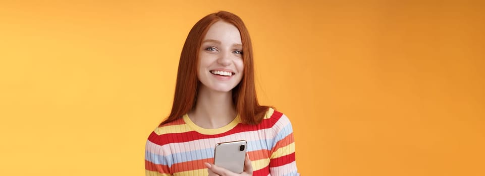 Friendly enthusiastic young redhead girl blue eyes using smartphone turn camera answer smiling broadly telling who sent message standing delighted orange background messaging, texting boyfriend.