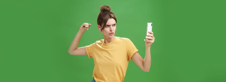 Woman making serious look to take selfie while listening music in wireless earphones posing and staring at smartphone screen daring and aggressive, liking post photos online over green background. Technology and lifestyle concept