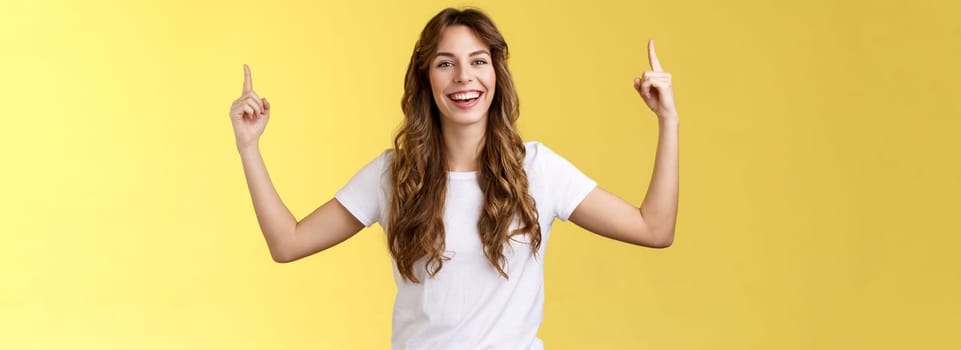 Cheerful amused happy lively young girl long curly haircut raise hands pointing up smiling toothy happily camera introduce excellent variant suggest you click site link advertising yellow background. Lifestyle.
